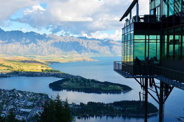Qué ver en Nueva Zelanda: 5 Lugares Imprescindibles para tu Viaje de Verano Moderna construcción de cristal junto a un lago de Nueva Zelanda, con una península y montañas de fondo, un lugar imprescindible para un viaje de verano.