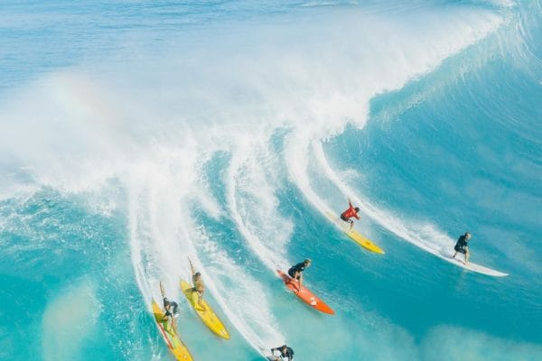 Hawái sin Mapa: Una Inmersión Privada en lo Esencial del Archipiélago Grupo de seis surfistas profesionales surfeando al mismo tiempo una imponente ola azul en la costa de Hawái.