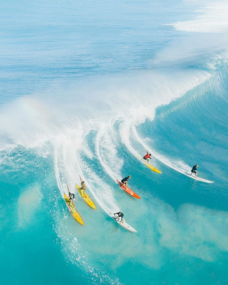 Grupo de seis surfistas profesionales surfeando al mismo tiempo una imponente ola azul en la costa de Hawái.