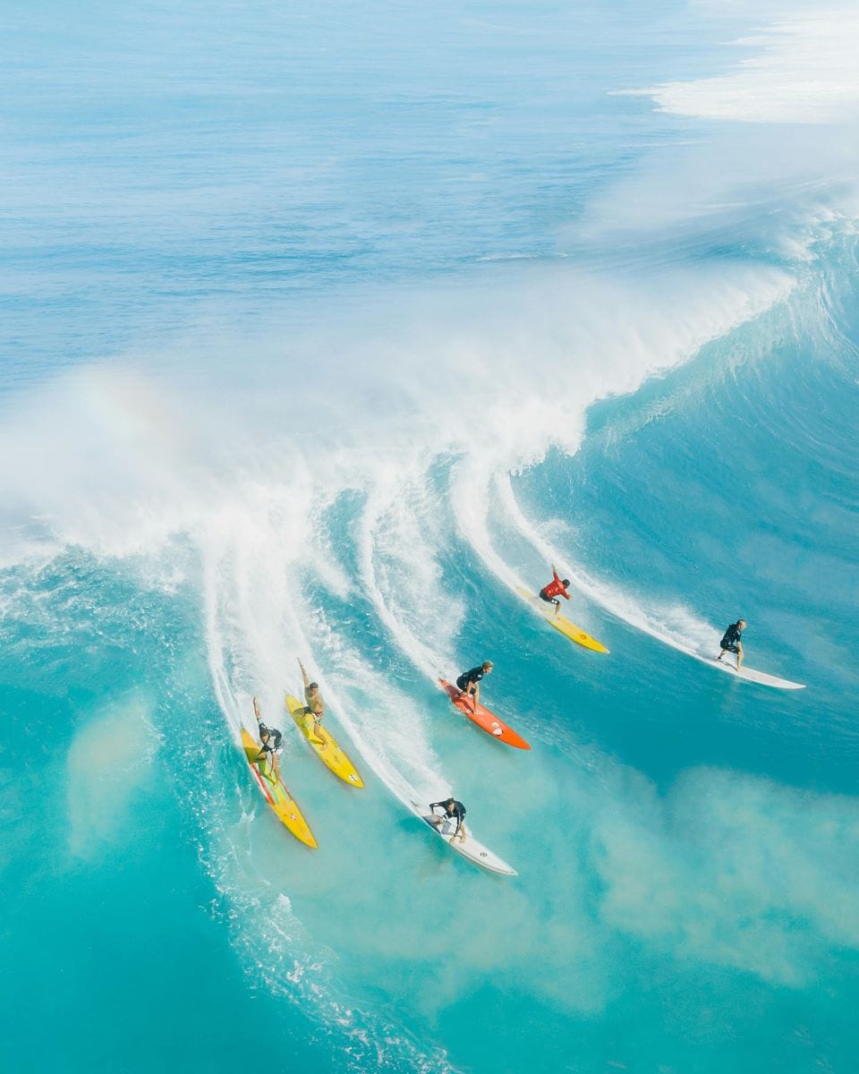 Grupo de seis surfistas profesionales surfeando al mismo tiempo una imponente ola azul en la costa de Hawái.
