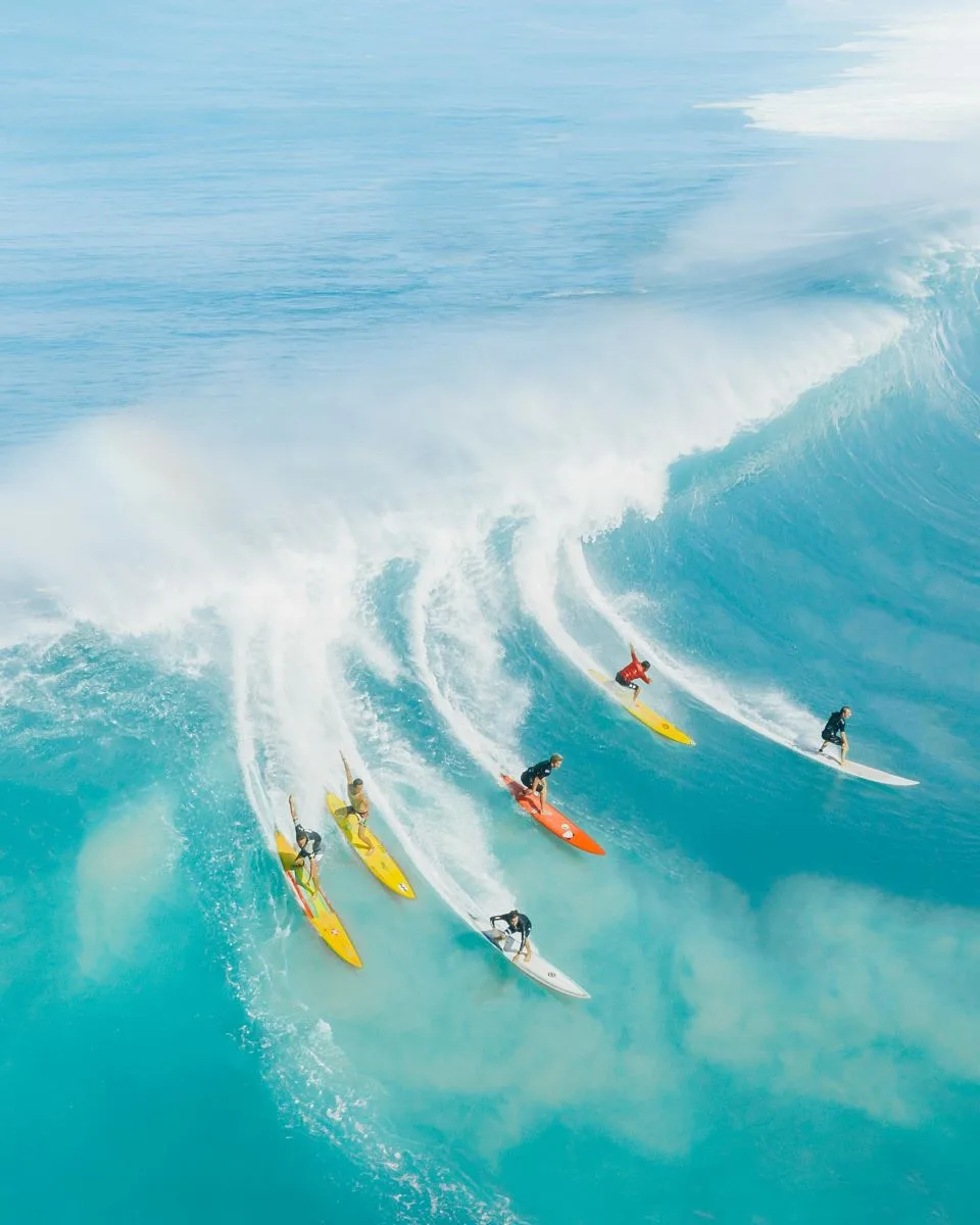 Grupo de surfistas profesionales descendiendo una ola gigantesca en Hawái, capturando la emoción de las experiencias privadas y extremas del archipiélago.