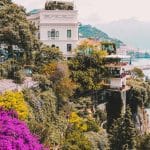 Los Secretos de los Jardines Privados de la Costa Amalfitana Flores multicolores en cascada desde un sendero de la Costa Amalfitana con vista al hotel Santa Caterina al fondo — ejemplo de belleza natural y alojamiento de lujo integrado al paisaje.