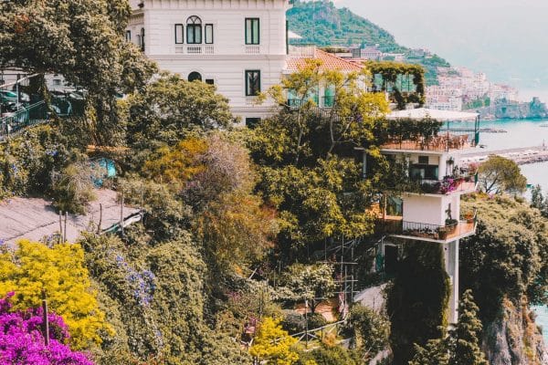 Los Secretos de los Jardines Privados de la Costa Amalfitana Flores multicolores en cascada desde un sendero de la Costa Amalfitana con vista al hotel Santa Caterina al fondo — ejemplo de belleza natural y alojamiento de lujo integrado al paisaje.