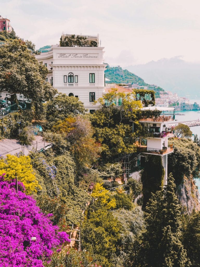 Los Secretos de los Jardines Privados de la Costa Amalfitana Flores multicolores en cascada desde un sendero de la Costa Amalfitana con vista al hotel Santa Caterina al fondo — ejemplo de belleza natural y alojamiento de lujo integrado al paisaje.