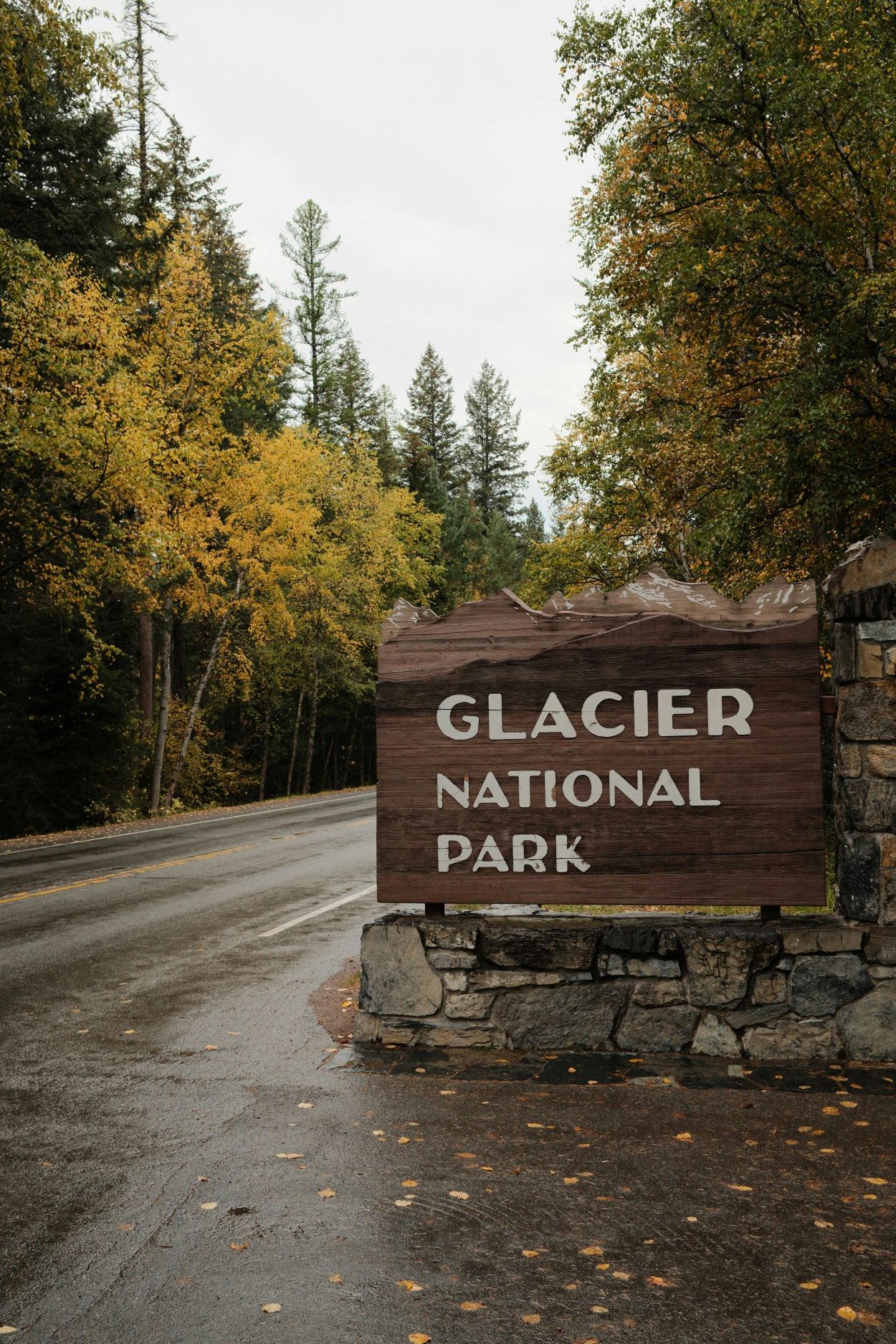 Cartel de madera en la entrada principal del Parque Nacional Glacier, Montana, con el nombre del parque tallado y bosques alpinos de fondo