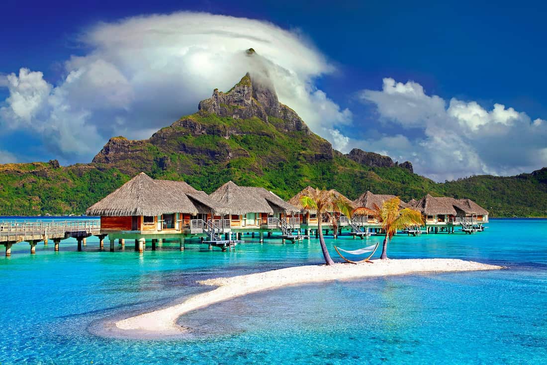 Bungalows de lujo sobre aguas turquesas frente a una isla de arena con hamaca y montañas de selva en la Polinesia Francesa.
