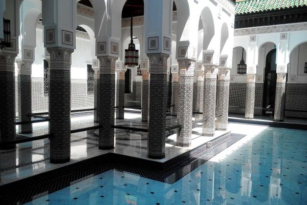 Patio interior de un riad de lujo en Marrakech con arcos árabes y columnas moriscas sobre espejo de agua y vegetación.