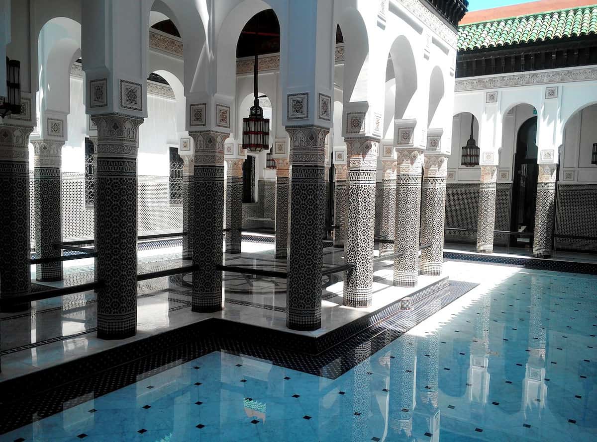 Patio interior de un riad de lujo en Marrakech con arcos árabes y columnas moriscas sobre espejo de agua y vegetación.