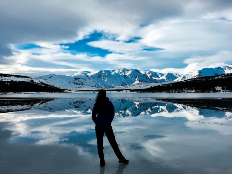 Silueta de mujer de espaldas mirando hacia una montaña nevada al borde de un lago helado en la Patagonia Argentina