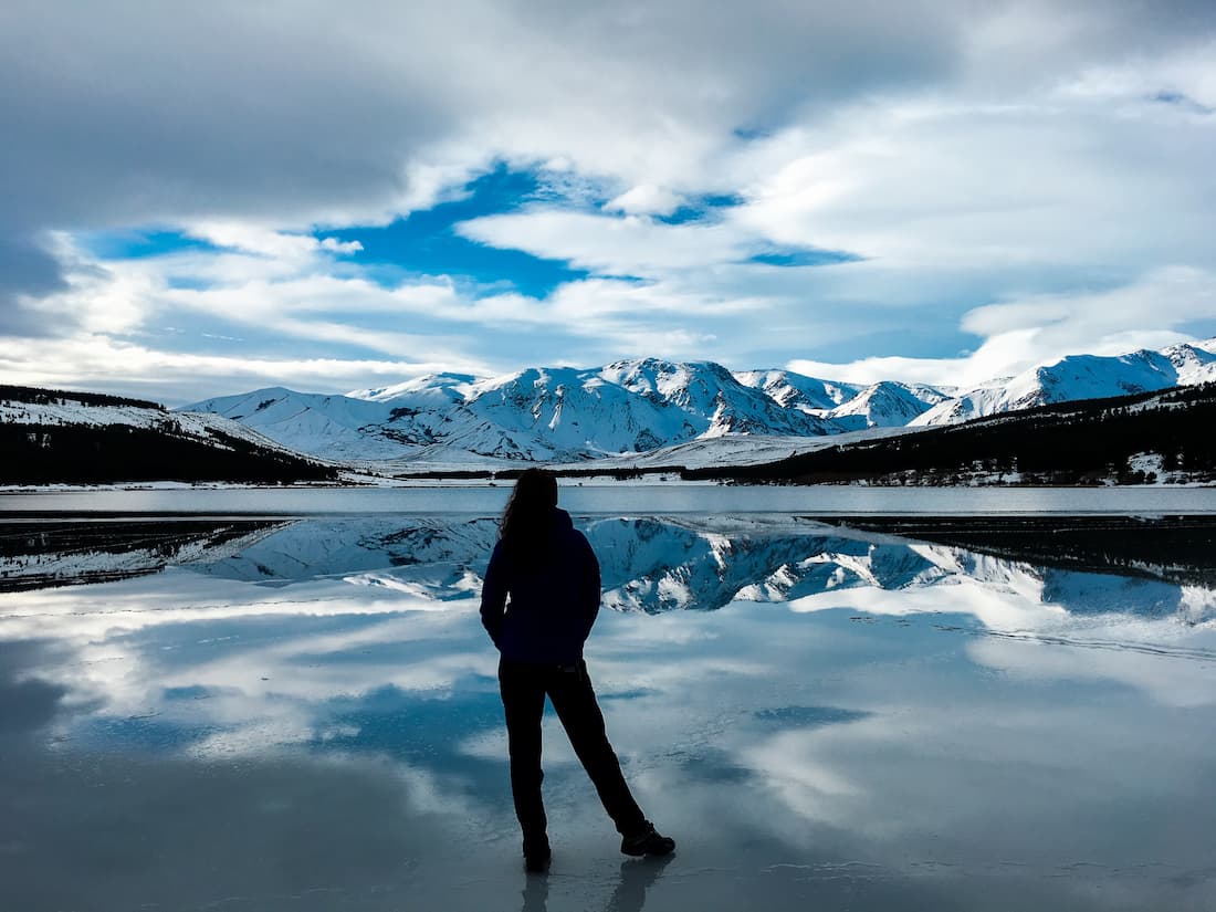 Silueta de mujer de espaldas mirando hacia una montaña nevada al borde de un lago helado en la Patagonia Argentina