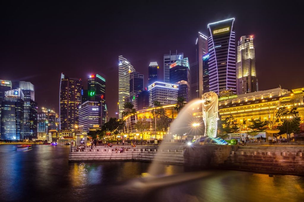 Merlion de Singapur con chorro de agua iluminado y el skyline nocturno de Marina Bay al fondo visto desde el mar