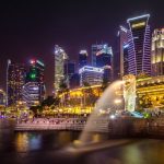 Merlion de Singapur con chorro de agua iluminado y el skyline nocturno de Marina Bay al fondo visto desde el mar