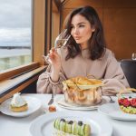 Mujer con copa de champagne sentada mirando por la ventana de un barco de crucero de lujo con postres en la mesa