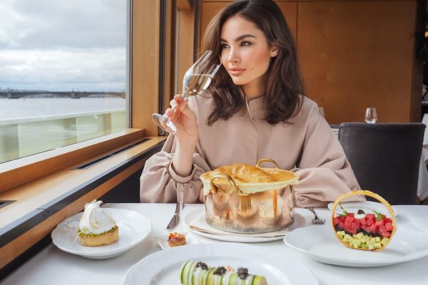Mujer con copa de champagne sentada mirando por la ventana de un barco de crucero de lujo con postres en la mesa