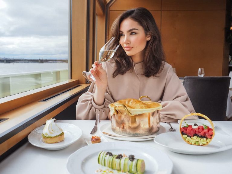 Mujer con copa de champagne sentada mirando por la ventana de un barco de crucero de lujo con postres en la mesa
