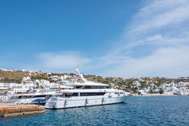 Cinco yates de lujo amarrados en un muelle en una bahía mediterránea con casas blancas sobre la colina al fondo