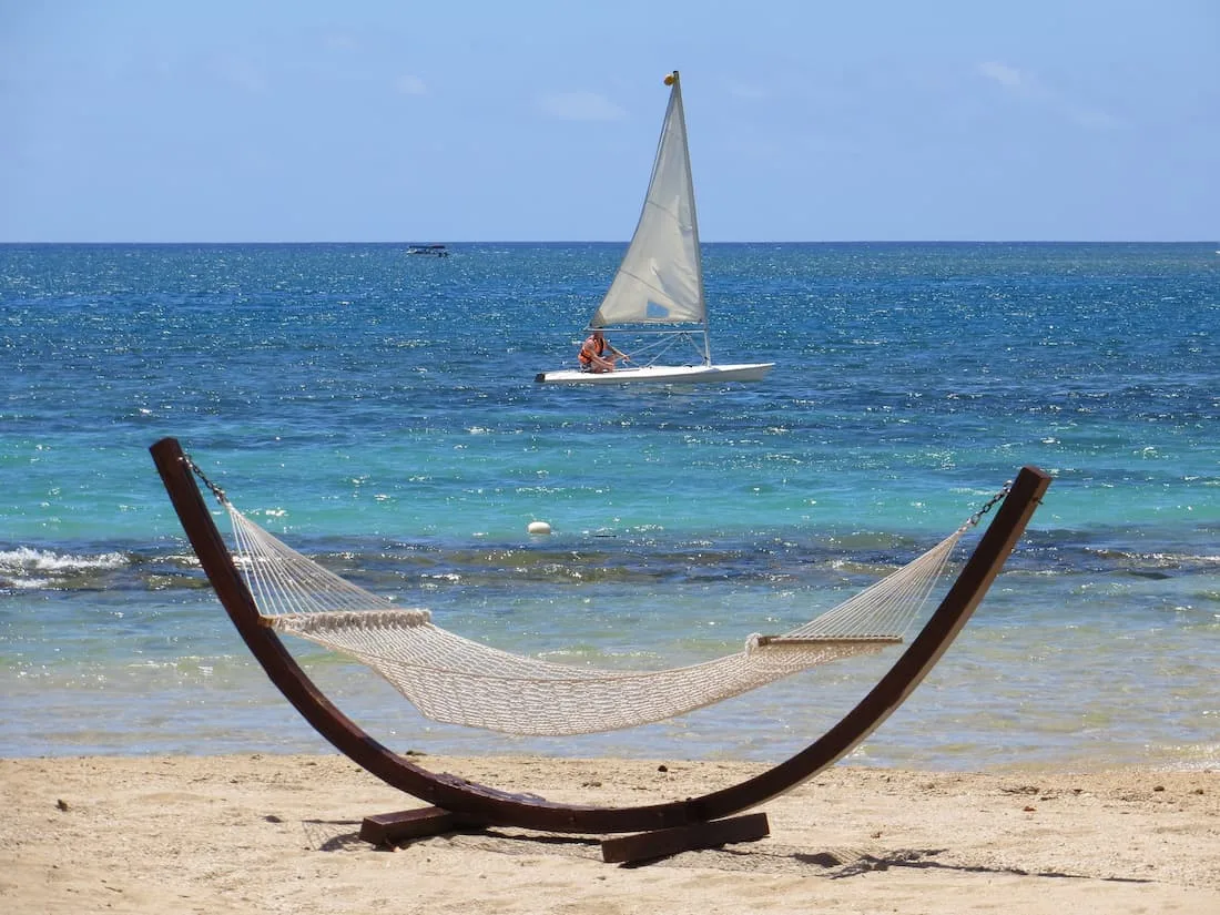 Hamaca en soporte de madera en la playa y chica sobre tabla de surf con vela en agua turquesa del Caribe