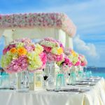 Mesa con mantel blanco, vajilla de lujo y decoración floral bajo pérgola blanca frente al mar para una luna de miel romántica