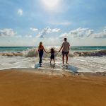 Pareja cogida de la mano con niño a la orilla del mar de espaldas a la cámara en vacaciones familiares.