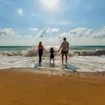 Pareja cogida de la mano con niño a la orilla del mar de espaldas a la cámara en vacaciones familiares.