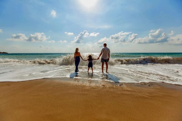 Pareja cogida de la mano con niño a la orilla del mar de espaldas a la cámara en vacaciones familiares.