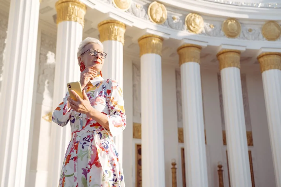Mujer elegante madura con vestido floral y gafas frente a arquitectura clásica con columnas doradas viajando sola después de los 50