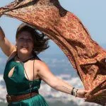 Mujer madura con vestido verde y pañuelo al viento en mirador con ciudad al fondo viajando sola después de los 50