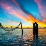 Pareja frente a frente tomados de la mano dentro del agua junto a una hamaca con atardecer de nubes de colores al fondo