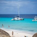Pequeña playa de Curaçao con personas entre sombrillas de paja y catamaranes anclados en mar transparente