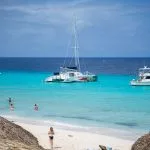 Pequeña playa de Curaçao con personas entre sombrillas de paja y catamaranes anclados en mar transparente