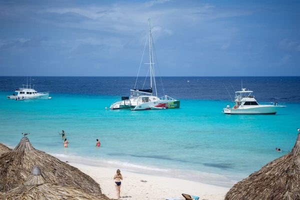 Pequeña playa de Curaçao con personas entre sombrillas de paja y catamaranes anclados en mar transparente