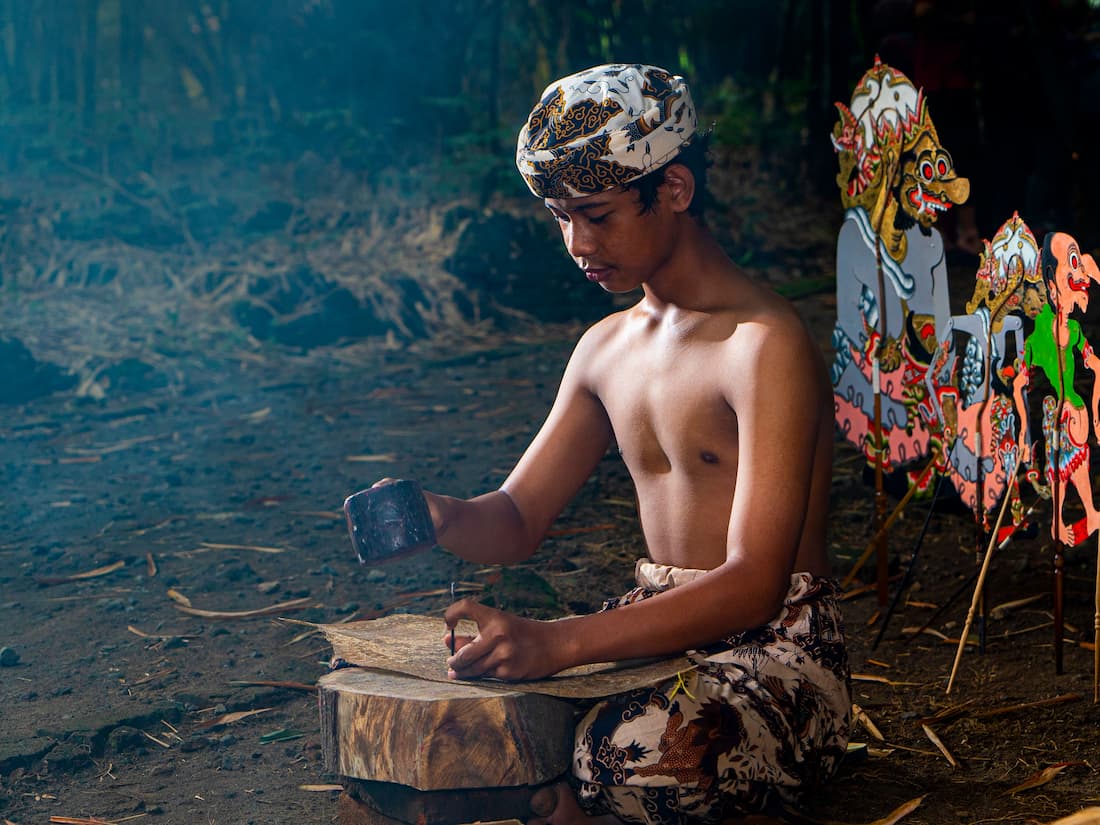 Joven artesano balinés tallando madera con herramientas tradicionales y figuras de wayang kulit al fondo en Ubud Bali