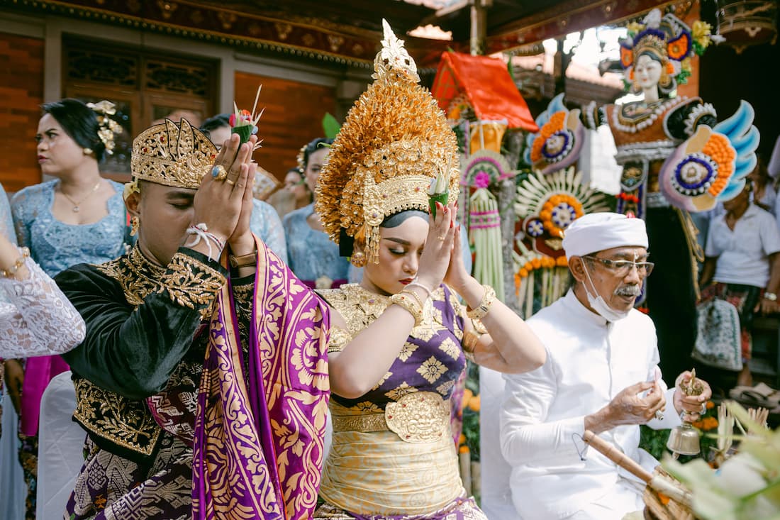 Pareja balinesa con trajes ceremoniales dorados y púrpura rezando durante ceremonia hindú en Ubud Bali Indonesia