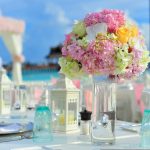 Mesa romántica decorada con flores hortensias rosas y linternas blancas con mar turquesa de Maldivas al fondo luna de miel de lujo