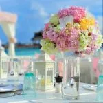 Mesa romántica decorada con flores hortensias rosas y linternas blancas con mar turquesa de Maldivas al fondo luna de miel de lujo