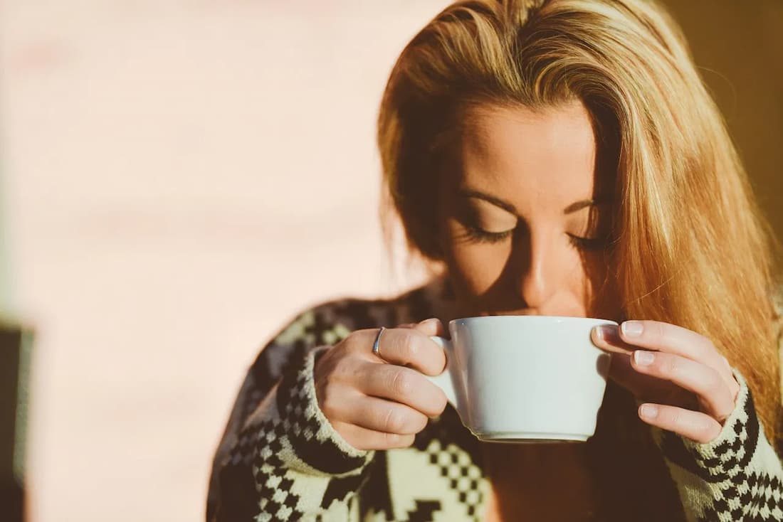 Mujer rubia con jersey de punto bebiendo café con las manos con luz cálida evocando el placer pausado del turismo lento