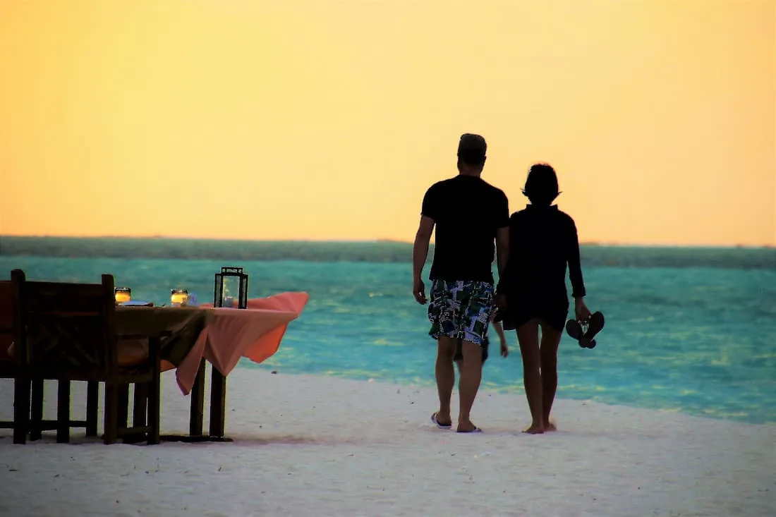 Pareja caminando descalza hacia el mar turquesa al atardecer junto a mesa de cena romántica con velas en playa de Maldivas