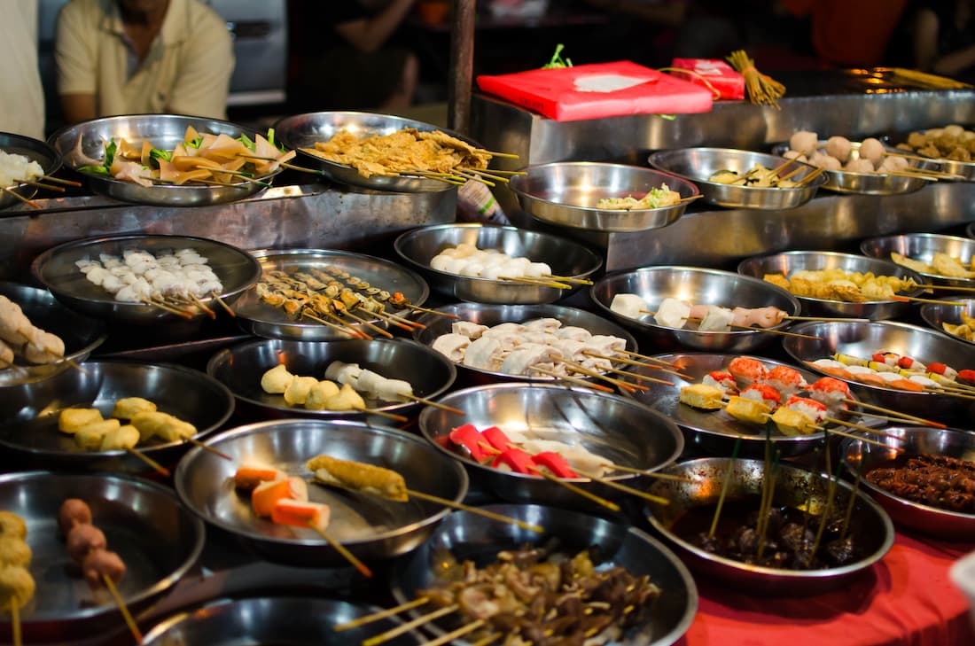 Puestos de gastronomía callejera con bandejas de metal y brochetas en hawker centre de Kuala Lumpur Malaysia